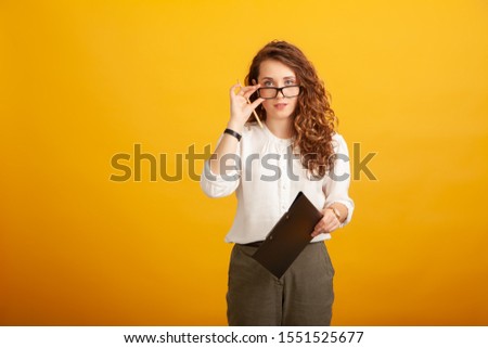 Beautiful young girl with a clipboard and a pencil puts down her glasses and looks at the camera standing isolated on yellow background
