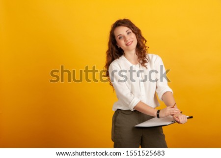 Beautiful young girl with a clipboard and a pencil in her hand smiles and looks at the camera standing isolated on yellow background