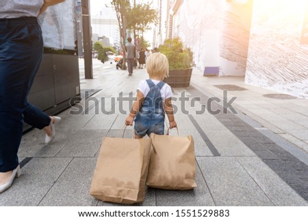 Small cute blond child is carring a lot of shopping bags at the street after black Friday sale. View from the back. Toddler is happy and feels she is a shopoholic. Royalty-Free Stock Photo #1551529883