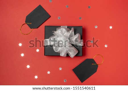Gift box wrapped in black paper with silver ribbon, stars confetti and price tags on bright red background. Copy space and top view. Black friday box gift present isolated.