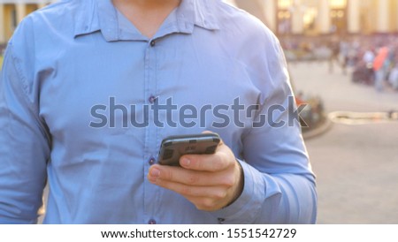 Hand of businessman in shirt standing among city street and browsing smartphone. Confident man writing message or using app on his phone during break in work. Blurred background