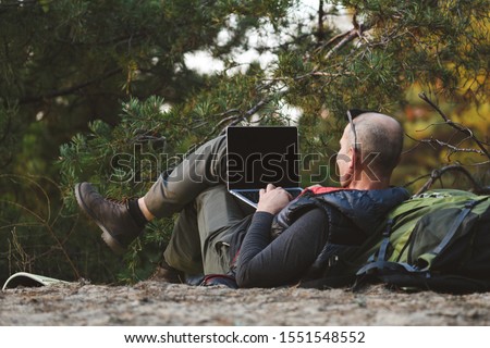 Elderly stylish man tourist use laptop in forest. Back view, male hiker lying on back leaning on backpack, with laptop computer, typing, blogging, browsing in forest. Freelance, distance work concept.