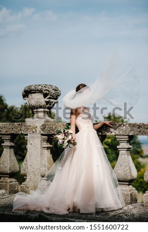 Young beautiful girl in elegant dress is standing and holding hand bouquet of pastel pink flowers and greens near old wall at nature. The wind throws a bridal veil outdoors.