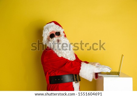Santa Claus with modern gadgets isolated on yellow studio background. Caucasian male model in traditional costume. Concept of Christmas, New Year's, winter mood, technologies. Using laptop, purchases.