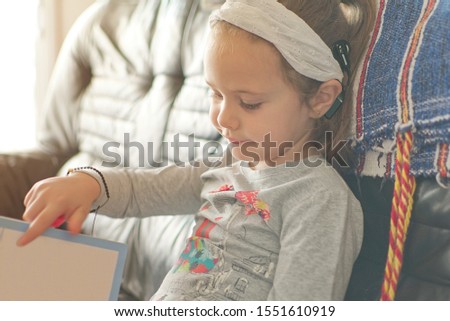 Blonde little girl with cochlear implant reading a book at home. Hear impairment and deaf community concept with empty copy space for Editor's text.