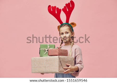 Portrait of little girl in deer antlers holding Christmas presents in her hands and smiling at camera isolated on pink background
