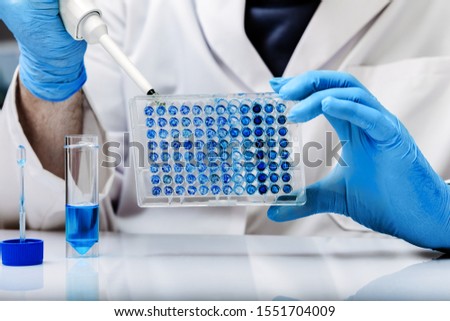 Biologist in lab holding a 96 well plate with biological samples for analysis / Scientist pipetting samples in micro plate in the laboratory Royalty-Free Stock Photo #1551704009