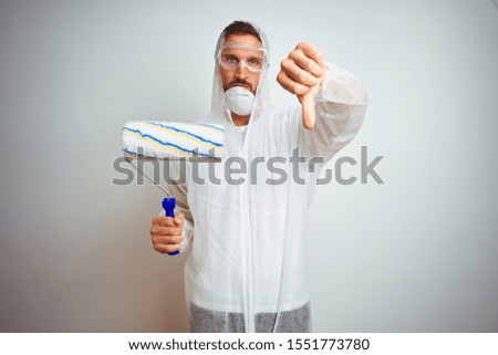 Painter man wearing professional worker equipment holding roller over isolated background with angry face, negative sign showing dislike with thumbs down, rejection concept