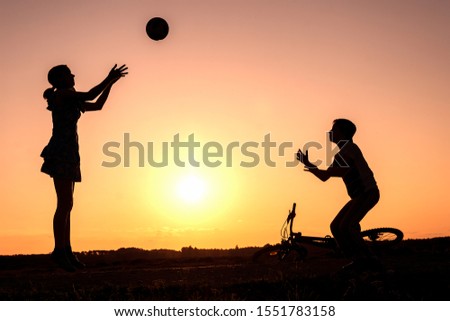 Brother with sister are playing with ball in nature, bicycle lies nearby,  silhouettes of playing children at sunset in countryside