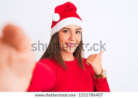 Girl wearing Christmas Santa hat make selfie by camera over isolated white background pointing and showing with thumb up to the side with happy face smiling