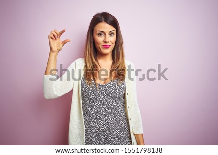 Young beautiful woman standing over pink isolated background smiling and confident gesturing with hand doing small size sign with fingers looking and the camera. Measure concept.