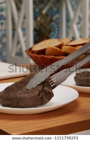Subject shot of a piece of bread fixed with a stainless steel tongs for cooking above a white plate. A wicker basket with biscuits is on the wooden work-surface against the ceramic tiles wall. 