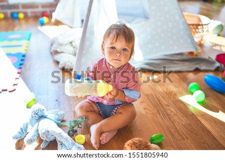 Adorable toddler holding feeding bottle around lots of toys at kindergarten