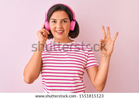 Young beautiful woman listening to music using headphones over isolated pink background doing ok sign with fingers, excellent symbol
