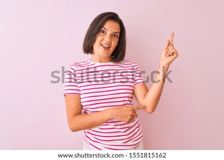 Young beautiful woman wearing striped t-shirt standing over isolated pink background with a big smile on face, pointing with hand finger to the side looking at the camera.