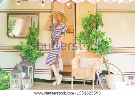 a cute woman with wavy blonde hair in a trendy summer striped dress on the background of a trailer camper van rests