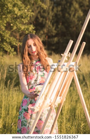 Young beautiful girl with long natural hair draws a picture while with an easel in nature. Girl enjoy solitude with nature.