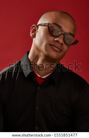Close-up shot of a young man with shaved head in a black shirt and with cat eyes retro sunglasses with a brown spotted half-frame and brownish lens. The photo is made on the red background.  