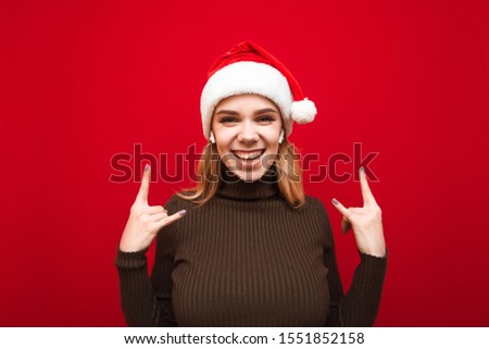 Portrait of a happy girl in a warm sweater and Santa hat listens to music in a cordless headset, looks into the camera, smiles and shows a heavy metal gesture. Christmas portrait. Isolated.
