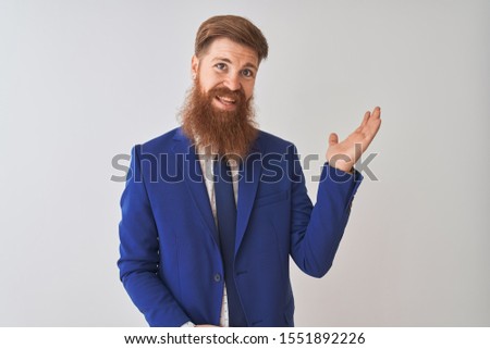 Young redhead irish businessman wearing suit standing over isolated white background smiling cheerful presenting and pointing with palm of hand looking at the camera.