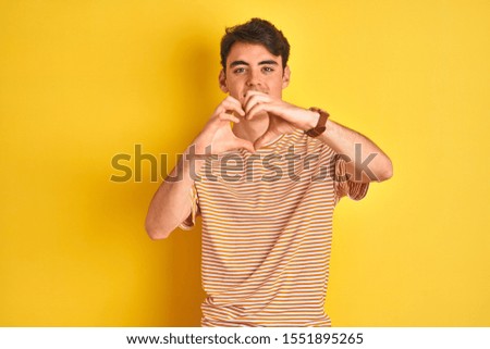 Teenager boy wearing yellow t-shirt over isolated background smiling in love doing heart symbol shape with hands. Romantic concept.