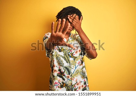 Afro man with dreadlocks on vacation wearing summer shirt over isolated yellow background covering eyes with hands and doing stop gesture with sad and fear expression. Embarrassed and negative concept