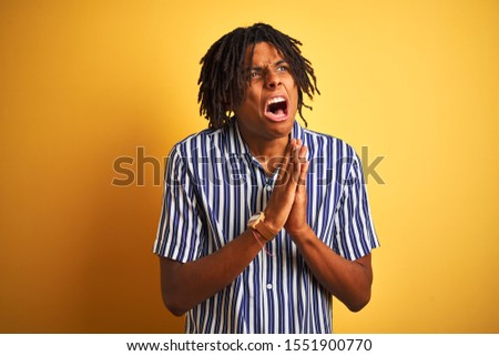 Afro man with dreadlocks wearing casual striped t-shirt over isolated yellow background begging and praying with hands together with hope expression on face very emotional and worried. Asking