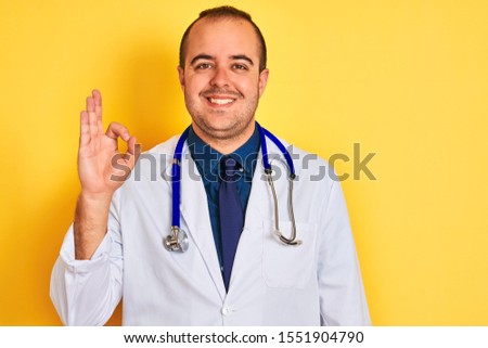 Young doctor man wearing coat and stethoscope standing over isolated yellow background smiling positive doing ok sign with hand and fingers. Successful expression.