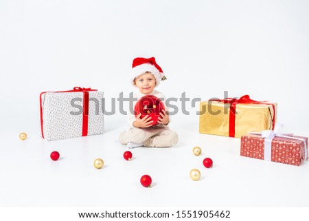 Little boy in Santas hat sitting between gifts and holding big red Christmas ball in hands. Isolated on white background. Holidays, christmas, new year, x-mas concept