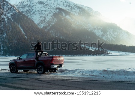 
Man sitting on the hood of a car. Hockey player looking for frozen pond. Shiny red pickup truck on ice covered road and snowy rural landscape. Offroad 4x4