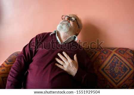 Old man with heart attack. Elderly senior man has chest pain. Unhappy man face in acute breast pain sitting on sofa at home, feel sick unwell. Sad aged man hand holding his heart. Heart attack symptom