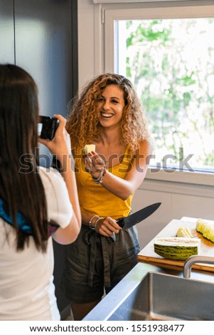 Vertical stock photo of a Two girls of different ethnicities in the kitchen, one shows a piece of melon to the camera of the other, which shoots a photo. Holidays and friends