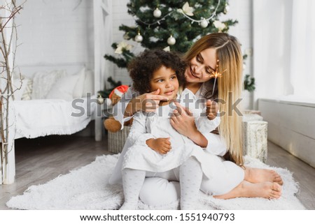 Merry Christmas and Happy Holidays! Cheerful mom and her cute daughter at Christmas tree. Parent and little child having fun near Christmas tree indoors. Loving family with presents in room.