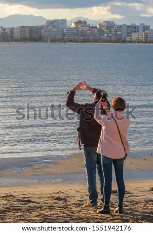 Close up of married couple takes amazing pictures of a beautiful sunset over the sea. The husband folded fingers in the shape of a heart on the sun background and the wife takes pictures on smartphone