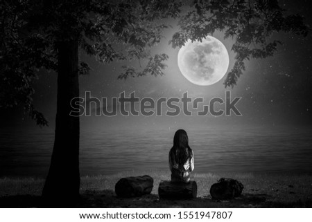 A black and white picture. The little girl sat and looked at the moon on a log by the sea on a full moon night beautifully.