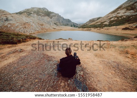 Stock aerial photo of a photographer girl from behind sitting in front of a lake surrounded by mountains with a camera in his hands. Travel and nature