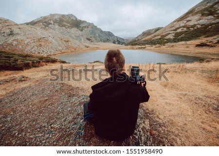 Stock photo of girl sitting in front of a lake surrounded by mountains with a camera in his hands. Nature and travel
