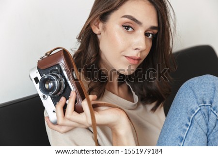Image of attractive young woman holding retro photo camera and sitting on sofa in apartment