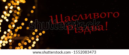 "Merry Christmas" inscription on Ukrainian language winter holidays concept post card picture with golden garland bokeh illumination on black background 