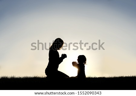 A silhouette of a young Christian mother teaching her little child to pray outside on a summer day.
