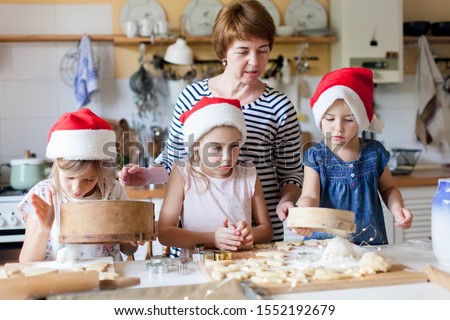 Family is cooking Christmas gingerbread cookies together in cozy home kitchen for dinner. Kids and young grandmother are baking holiday meal. Girls help woman. Lifestyle moment. Children chef concept
