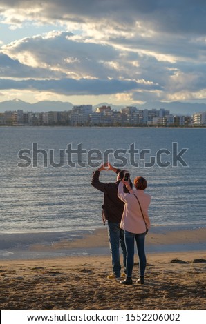 A married couple takes amazing pictures of a beautiful sunset over the sea. The husband folded fingers in the shape of a heart on the sun background and the wife takes pictures on smartphone. 