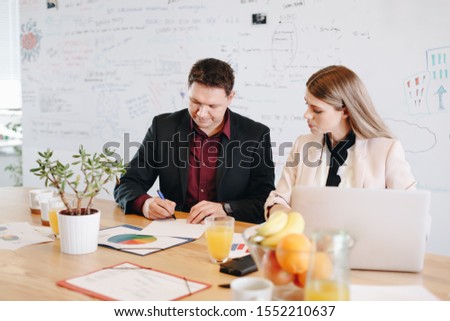 Businessman and businesswoman sitting at table and signing finalized documents in  modern open office. Candid moment, man becoming partner in law firm.