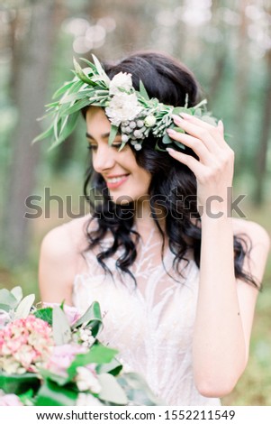 Beautiful smiling happy brunette bride with stylish bouquet and wreath on the background of green forest