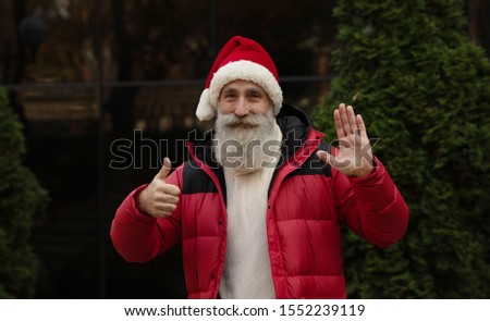 Senior man with beard in red santa hat presenting something outdoor. Xmas. New Year.