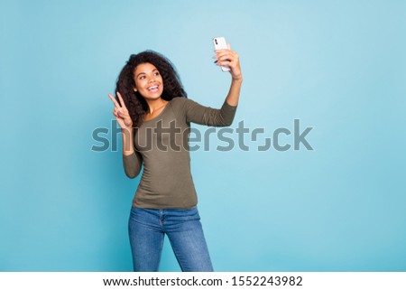 Portrait of positive cheerful wavy hair afro american girl blogger influencer have weekend journey take selfie on mobile phone make v-sign wear green sweater denim jeans isolated blue color background