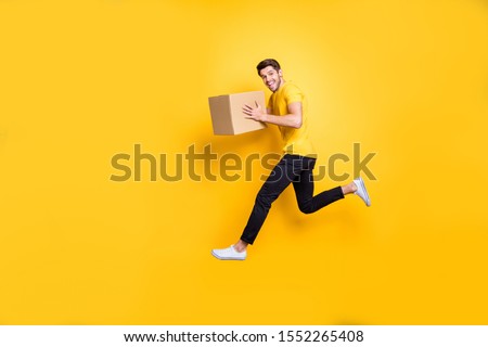 Full size photo of handsome guy jumping high holding parcel box hurry move new apartments transportation wear casual t-shirt pants isolated yellow color background