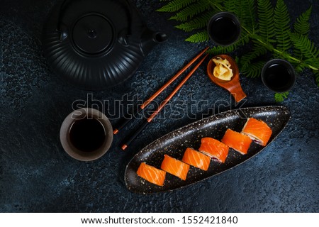 A set of sushi rolls Philadelphia with red fish and cream cheese lies in a plate boat. Sushi rolls on a black background with a teapot of Chinese tea. Top view