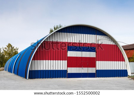 Close-up of the national flag of Costa Rica
 painted on the metal wall of a large warehouse the closed territory against blue sky. The concept of storage of goods, entry to a closed area, logistics