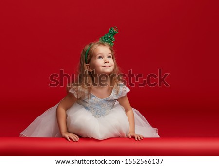 Cute little child girl in Christmas hat and fashion white princess dress and hoop on the head in the shape of Christmas trees having fun on red background. Empty space for text. Banner 